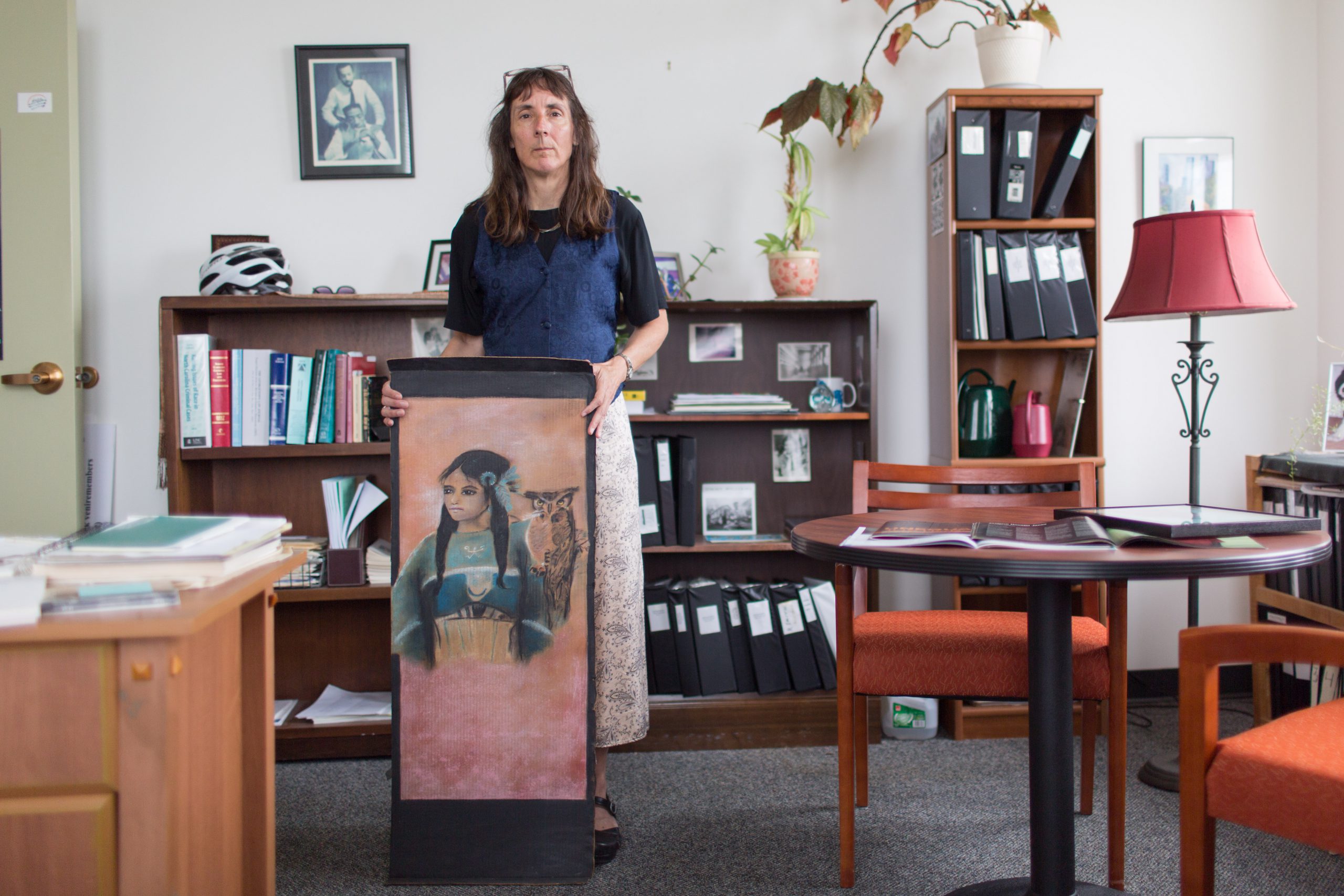Center for Death Penalty Litigation attorney and executive director Gretchen Engel in her office, holding a large-scale painting of a Native American girl in blue dress with an owl perched near her shoulder.