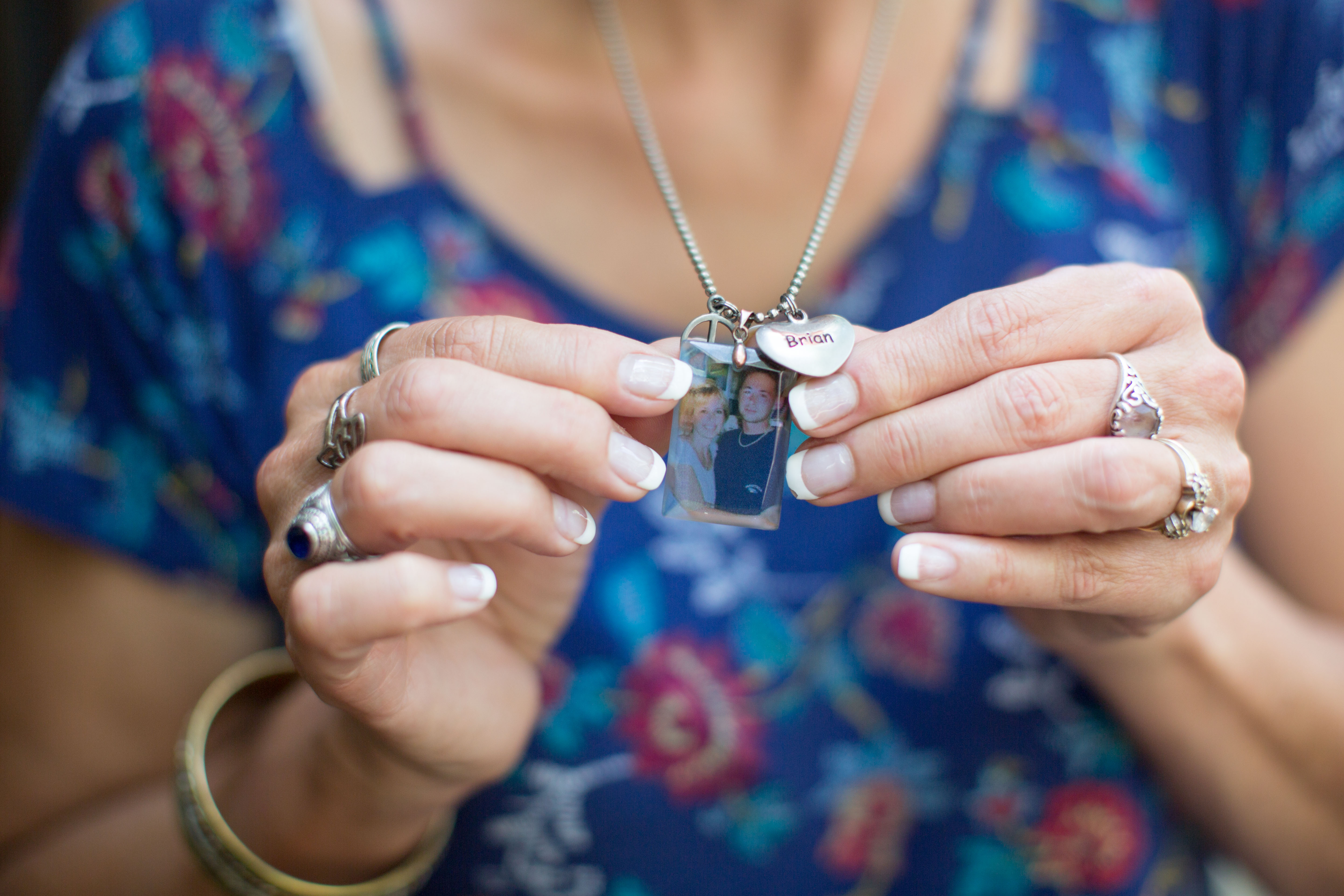 Lynda holds a her necklace toward the camera. On it, there's a photograph of her and her son Brian, along with a small heart trinket that shares his name. In the photo, her