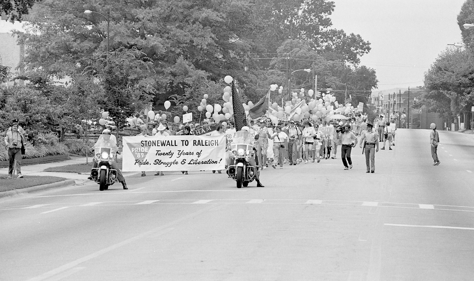 A black-and-white photo of a 1989 LGBTQ+ pride march in Raleigh, North Carolina. Marchers hold a banner reading “Stonewall to Raleigh: Twenty Years of Pride, Struggle & Liberation!” surrounded by balloons and signs, flanked by motorcycles.